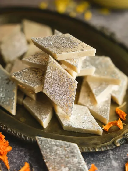 A clean, simple shot of Kaju Katli on a traditional brass plate, garnished with marigold petals. This image focuses on the texture and classic diamond shape of the sweet.