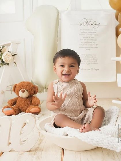 A huge, happy smile from the birthday boy during his neutral-toned first birthday shoot. He's sitting in a bowl, surrounded by classic toys and balloons.
