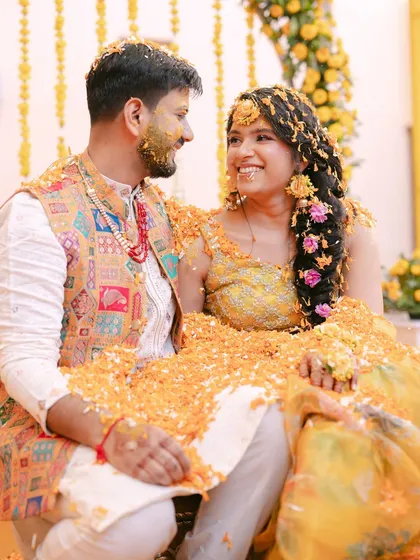 A sweet, candid moment between the couple during their Haldi ceremony, covered in marigold petals and sharing a happy, loving gaze.