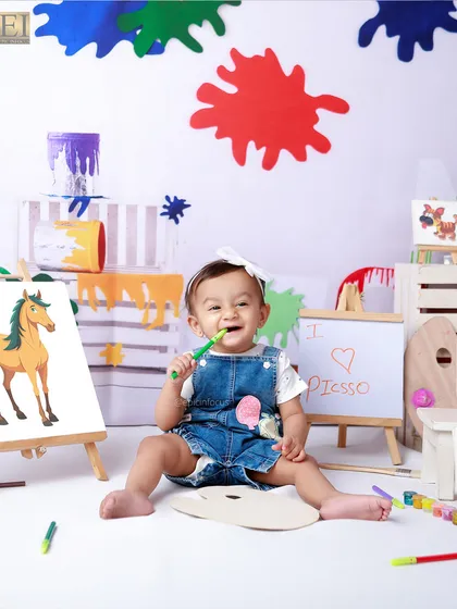 A happy baby artist, dressed in a denim jumper, chews on a paintbrush in our colorful studio. This theme is all about embracing mess and creativity.