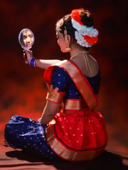 A creative shot from the back, showing the dancer's reflection. This perspective highlights the traditional braided hair adorned with flowers and the intricate design of the blouse.
