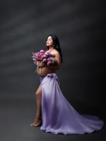 A moment of quiet beauty. The expectant mother holds a bouquet of purple flowers, her lavender satin wrap dress adding a touch of elegance and softness.