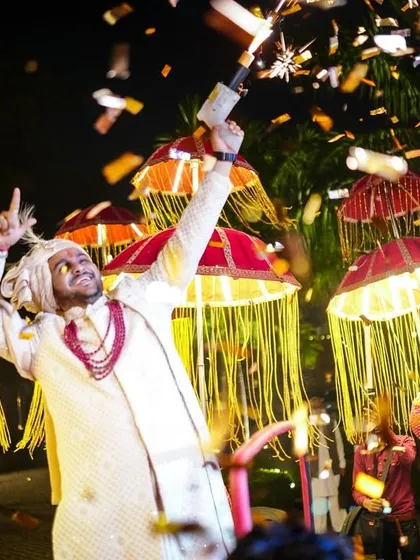 The groom's celebratory moment during the baraat, with a confetti cannon and traditional umbrellas.