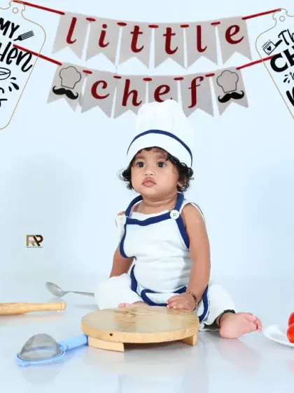 A baby dressed as a "Little Chef" sits in a clean, white kitchen setup. The chef's hat, apron, and miniature cooking props make for an adorable and creative photoshoot.