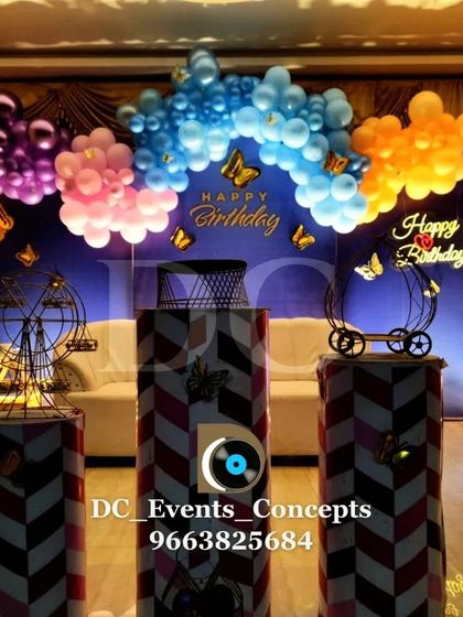 A whimsical butterfly-themed birthday party setup. The multi-colored balloon arch and decorative props like miniature ferris wheels create a magical and playful atmosphere.