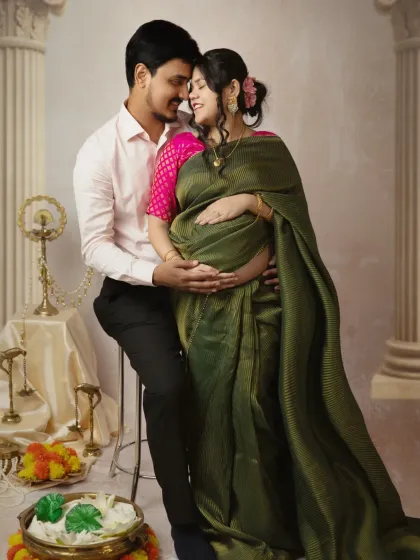 An intimate pose where the couple embraces, both cradling the baby bump. The mother looks radiant in an olive green saree, and the traditional props in the background add a sense of heritage.