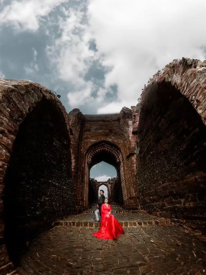 A dramatic wide-angle shot taken from within a brick archway, looking out at the couple. The bride's vibrant red dress provides a stunning focal point against the rustic textures and cloudy sky.