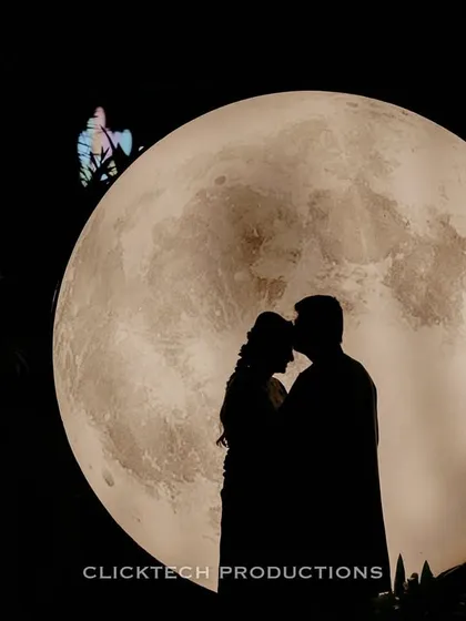 A creative and romantic silhouette of a couple against a giant, illuminated moon prop during a night pre-wedding shoot, demonstrating a dramatic and artistic photography style.