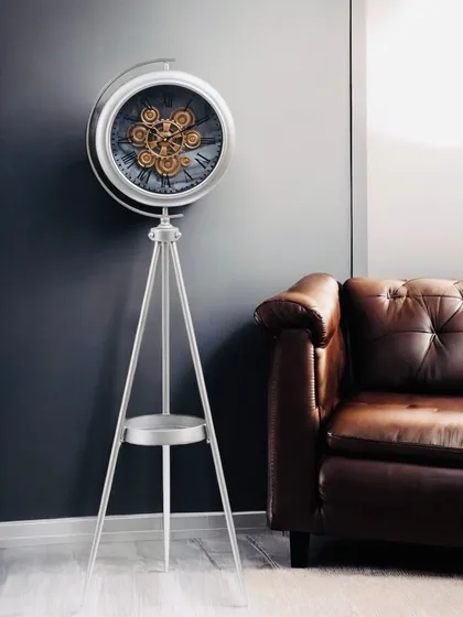 A luxury tripod standing clock crafted from high-quality steel with a silver metallic finish. The exposed moving gears and functional lower shelf make it a timeless piece of decor.