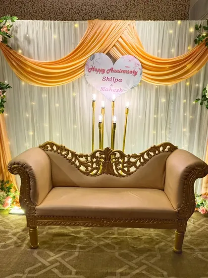 An elegant and intimate seating arrangement for a wedding anniversary celebration. The decor includes a beautiful backdrop with white and peach fabric draping, fairy lights, and floral arrangements in the corners.