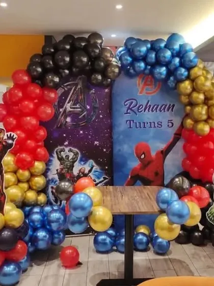 A superhero-themed birthday party featuring an Avengers backdrop. The decor includes balloon columns with Iron Man and Hulk cutouts.