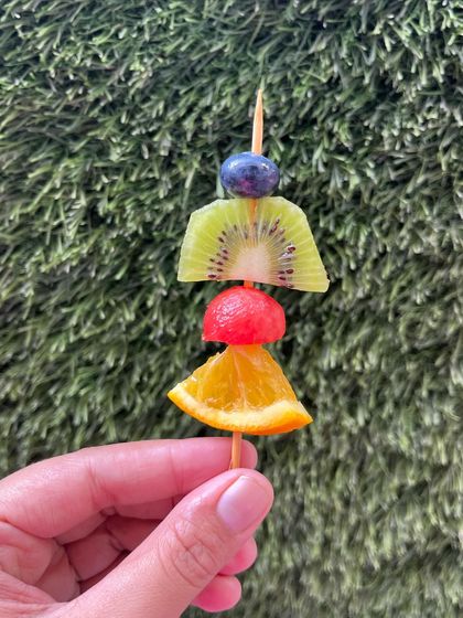 A single, vibrant fruit skewer featuring a slice of orange, watermelon, kiwi, and a blueberry. It's a burst of freshness in every single bite.