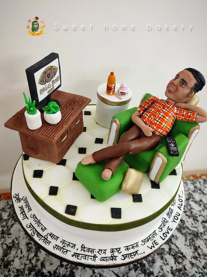 This cake was made for a dad's birthday and retirement party, celebrating his love for watching Bigg Boss. It features a detailed figurine of him relaxing in his favorite chair, complete with a TV, remote control, and his favorite snacks.