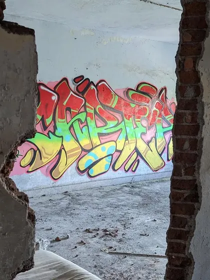 The same piece viewed through a hole in a broken wall. This framing gives a unique perspective on how graffiti exists within abandoned and forgotten spaces.