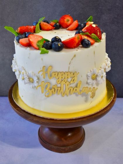 A beautiful birthday cake topped with a crown of fresh berries. This simple yet elegant design features fresh strawberries and blueberries, delicate white daisies, and a gold "Happy Birthday" script.