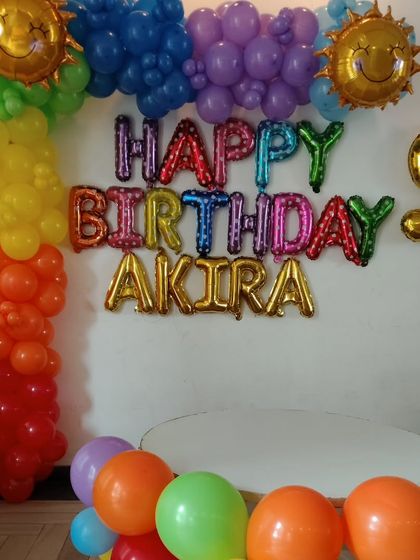 A vibrant rainbow-themed decoration for a 9th birthday. This setup includes a colorful balloon column, rainbow "Happy Birthday" foil letters, and fun smiling sun foil balloons.