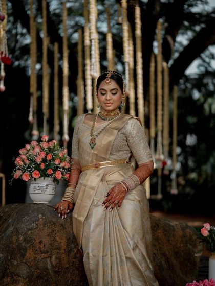 A serene portrait of a bride seated outdoors. The natural setting provides a beautiful contrast to the opulence of her golden Kanchipuram saree and custom blouse.