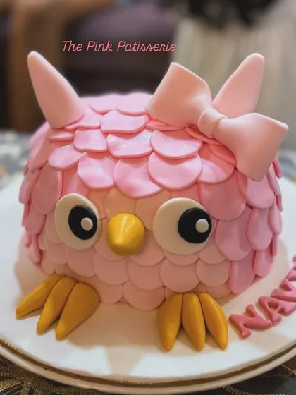 A cute pink owl cake for Navya. The cake is covered in individual fondant "feathers" and features big, expressive eyes and a bow.