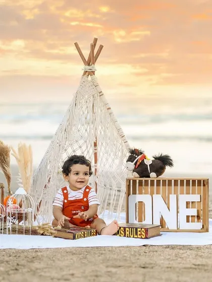 A happy one-year-old enjoying a boho-themed picnic setup on the beach at sunset.