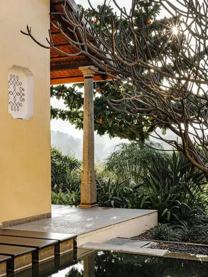 A close-up of the traditional stone pillar and decorative window at Dvara Resorts. The morning sun backlights the frangipani tree, creating a warm and ethereal glow.