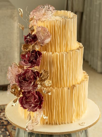 A magnificent three-tier cake in a warm butter yellow, completely covered in vertical wafer paper ruffles. A cascade of sugar flowers in deep burgundy and soft pink adds a dramatic and beautiful contrast.