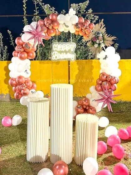 An elegant outdoor birthday setup featuring a balloon arch in rose gold and white. The arrangement includes star-shaped foil balloons and fluted pedestals for a sophisticated look.