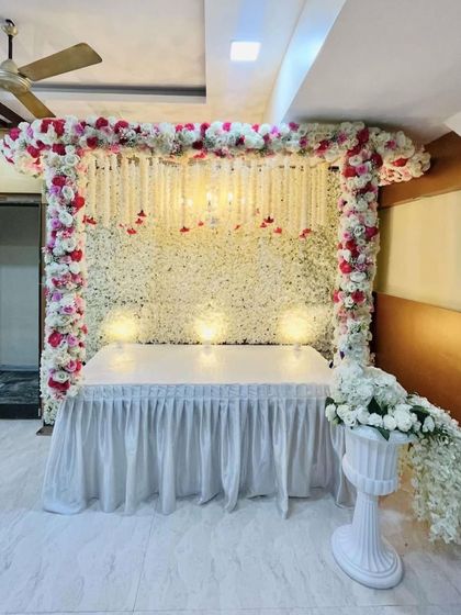 A beautiful backdrop made of a dense wall of white flowers with hanging red flower strings. The frame is decorated with pink and white roses, creating a soft and elegant look.