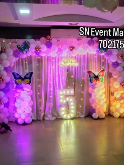A butterfly-themed stage decoration with dramatic lighting. The purple and pink lights enhance the balloon pillars and backdrop, creating a beautiful ambiance for a 5th birthday party.