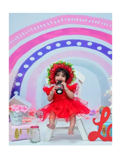 Dressed in a vibrant red dress with a floral bonnet, this baby girl playfully chews on a prop. The colorful rainbow background makes this a cheerful and eye-catching portrait.