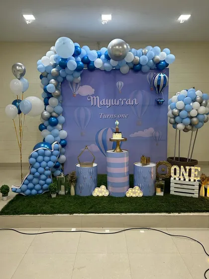 A full view of the hot air balloon theme stage. This angle shows the arrangement of plinths for the cake, the light-up number '1', and the artificial grass base that completes the look.
