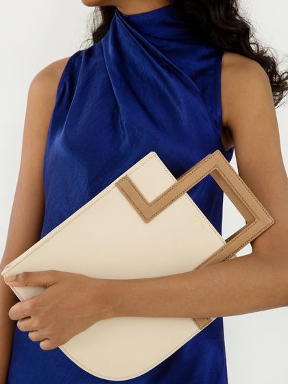 A clean, minimalist shot showcasing a cream and tan handbag. The bag is styled with a simple blue dress to make its unique design and color palette stand out.