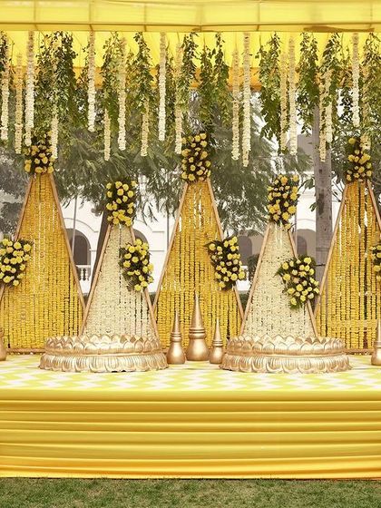 A traditional Haldi stage design with a modern twist. The backdrop features triangular panels covered in marigolds, with hanging floral strings and elegant gold finials.