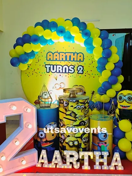 Everyone loves Minions! This fun setup for a second birthday includes a custom Minion-themed backdrop, character-printed plinths for the cake, a large light-up number '2', and of course, a blue and yellow balloon arch.