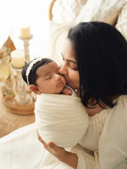 A close-up of a mother kissing her swaddled newborn's cheek. The soft, warm lighting and boho decor in the background add to the gentle mood of the photo.