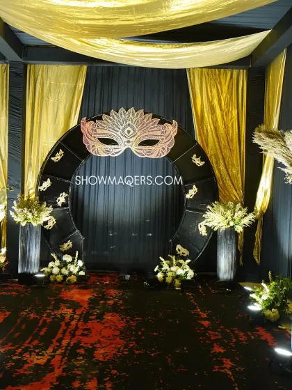 A grand photo arch designed for the masquerade ball theme. The circular structure is topped with a large, ornate mask, framed by black and gold drapes and floral arrangements.