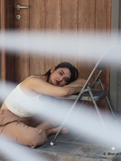 An introspective portrait of a model in a casual white top and tan pants, seated indoors. The composition, shot through a grate, adds a layer of artistic framing.