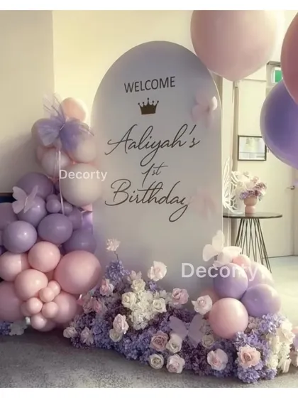 A personalized welcome sign for Aaliyah's first birthday, framed by pastel pink and purple balloons and delicate flowers for a soft, elegant look.
