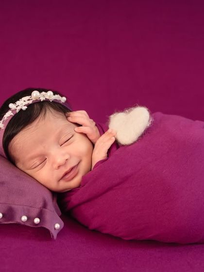 A heart-melting smile from a newborn dreaming sweet dreams, wrapped snugly in a purple swaddle with a tiny heart detail.