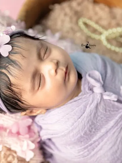 A close-up of a sleeping newborn's face, showcasing her delicate features and peaceful expression. The soft lavender wrap and floral headband complete this angelic look.