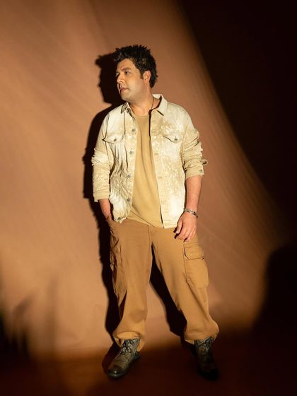 A full-length portrait of actor Varun Sharma. The lighting is designed to create a spotlight effect against the brown background.