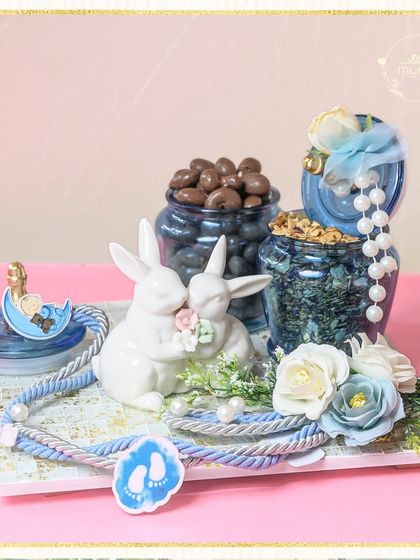 This charming baby boy hamper is filled with adorable details, including two kissing bunny figurines, jars of chocolate treats, and blue floral accents, all tied together with a nautical rope.