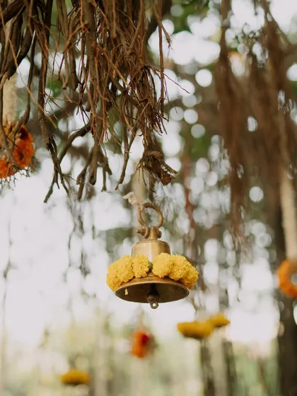 Attention to detail is key. Here, a traditional brass temple bell is adorned with fresh marigolds, hanging from the natural roots of a banyan tree to create a rustic and sacred ambiance.