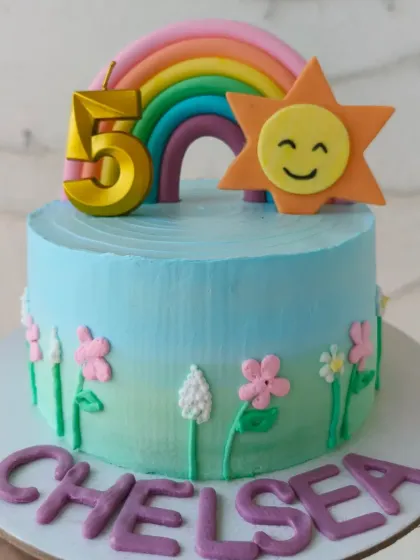 A cheerful rainbow themed cake with a smiling sun and fondant flowers.