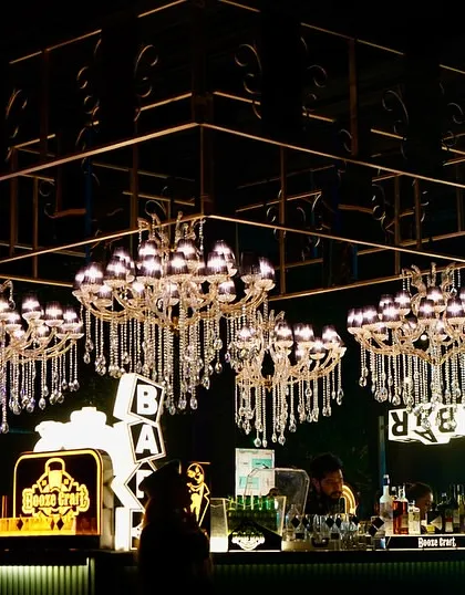 A wide shot of the glamorous bar at a 25th-anniversary party. The combination of crystal chandeliers, modern light fixtures, and a well-stocked bar creates a sophisticated and inviting atmosphere.