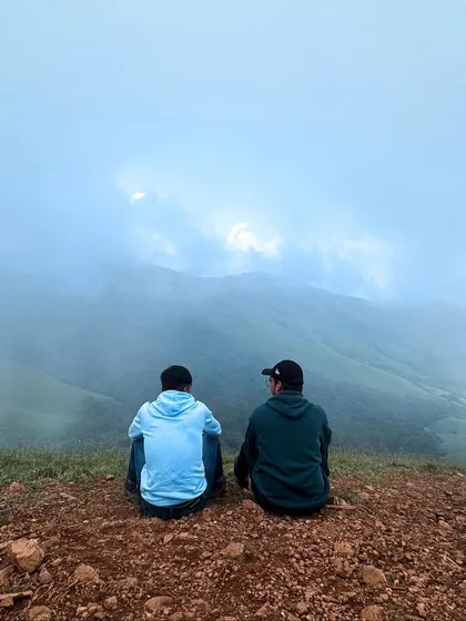 Two friends sitting at the edge of Netravathi peak, looking out at the mist-filled valley below.