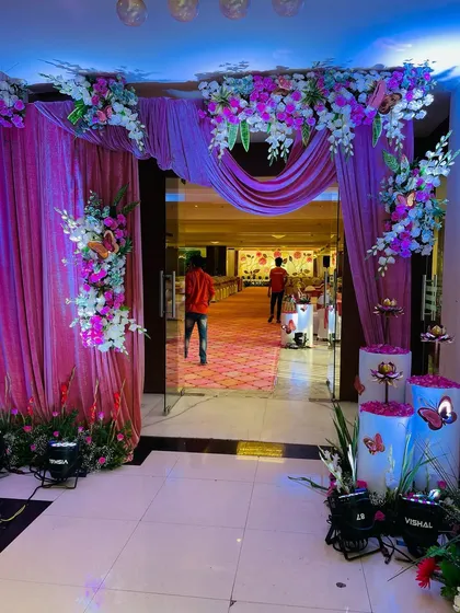 The entrance to a naming ceremony hall, decorated with pink drapes, floral arrangements, and illuminated pillars with butterfly accents.