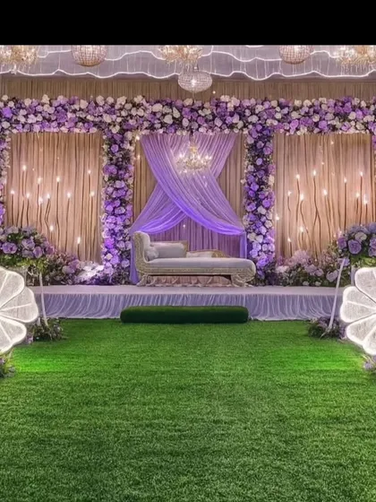 A grand and elegant stage designed for a reception or ring ceremony. The decor features a beautiful lavender and white floral arrangement, layered fabric draping with fairy lights, and large illuminated floral props, creating a magical ambiance for your special event.