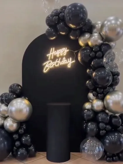 A sleek and modern birthday setup with a black arched backdrop. The decoration features a black and silver balloon garland and a "Happy Birthday" neon sign.