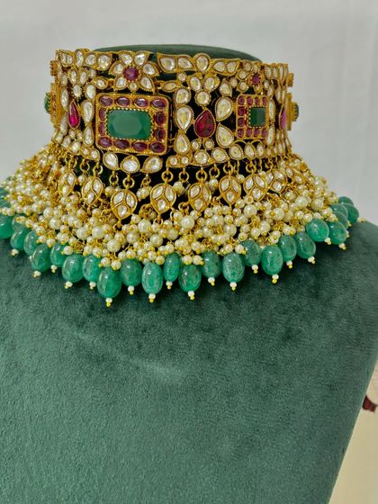 A close-up of a heavy bridal choker with green beads and pearl detailing. The combination of red and green stones makes it a traditional rental choice.