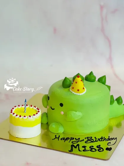 An adorable dinosaur-shaped cake for a birthday celebration. This cute green dino comes with its own mini birthday cake, making it a fun and unique design.